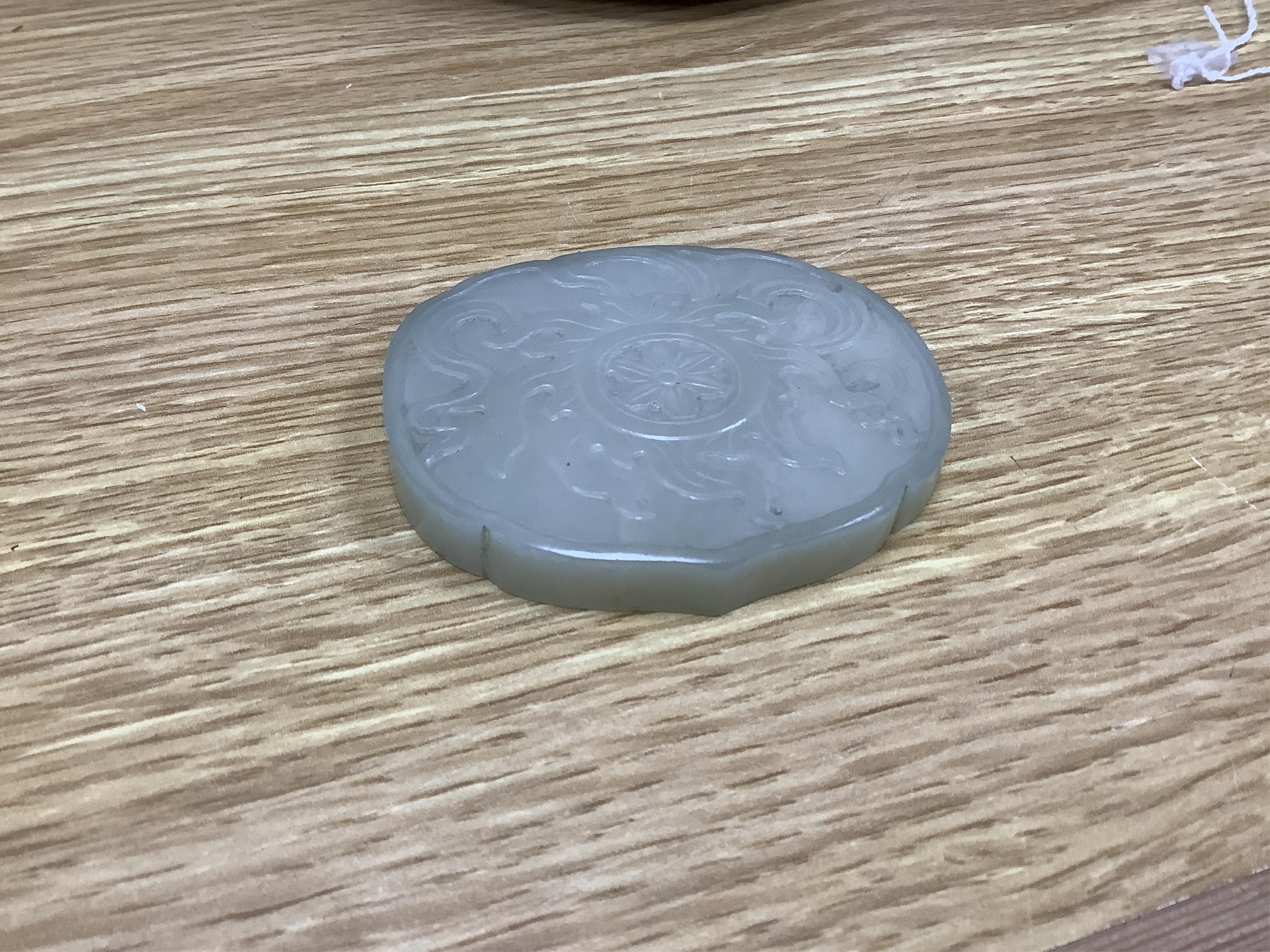 A Chinese pale celadon jade cover for a box, 19th century, carved in relief with a flaming Buddhist wheel emblem, 7.5cm wide. Condition - box base lacking, cover with feint crack